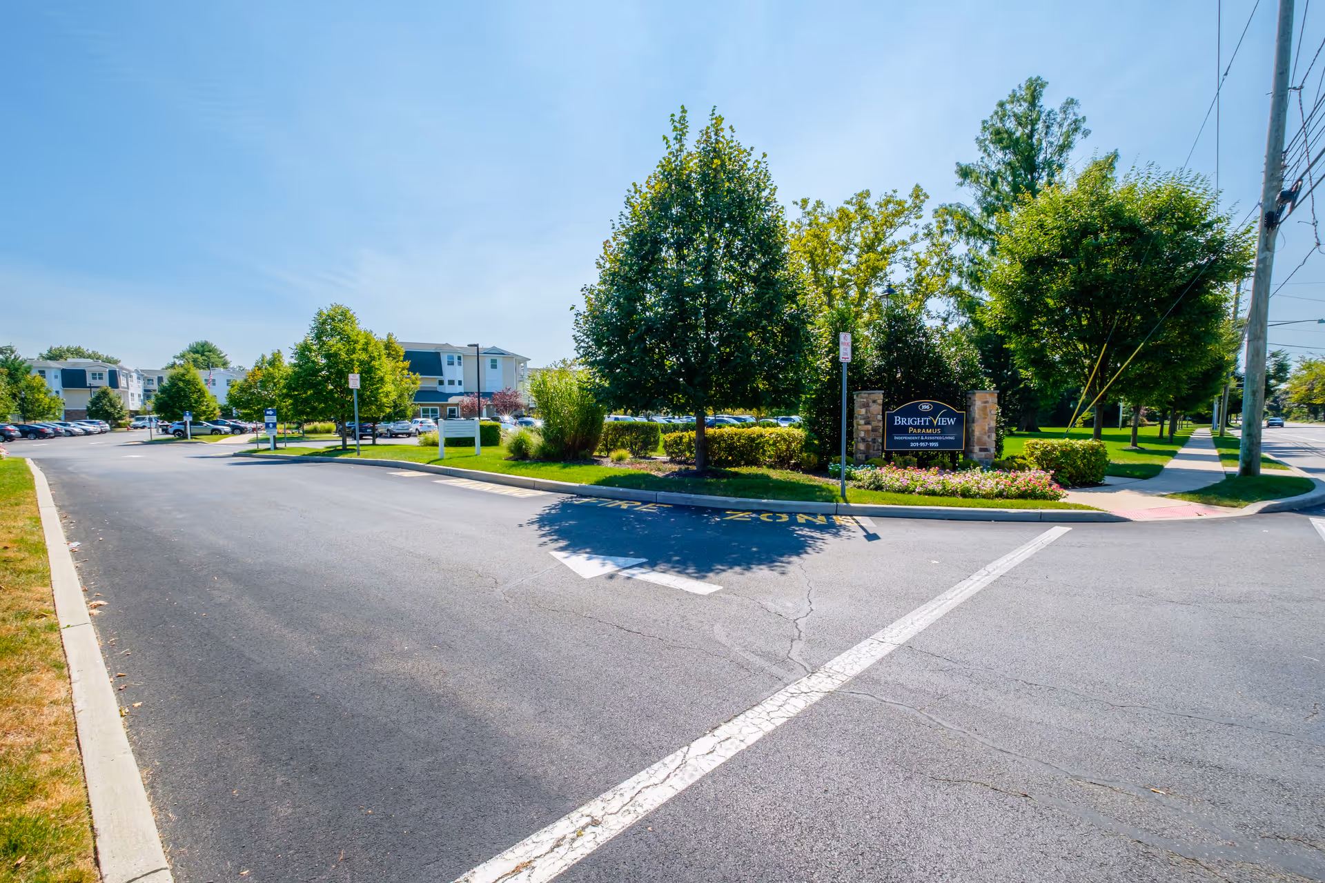View of the entrance road to Brightview Paramus senior living facility with a landscaped area featuring trees and flowers. The facility's sign is visible on the right side, surrounded by greenery, with apartment buildings and parked cars in the background under a clear blue sky.