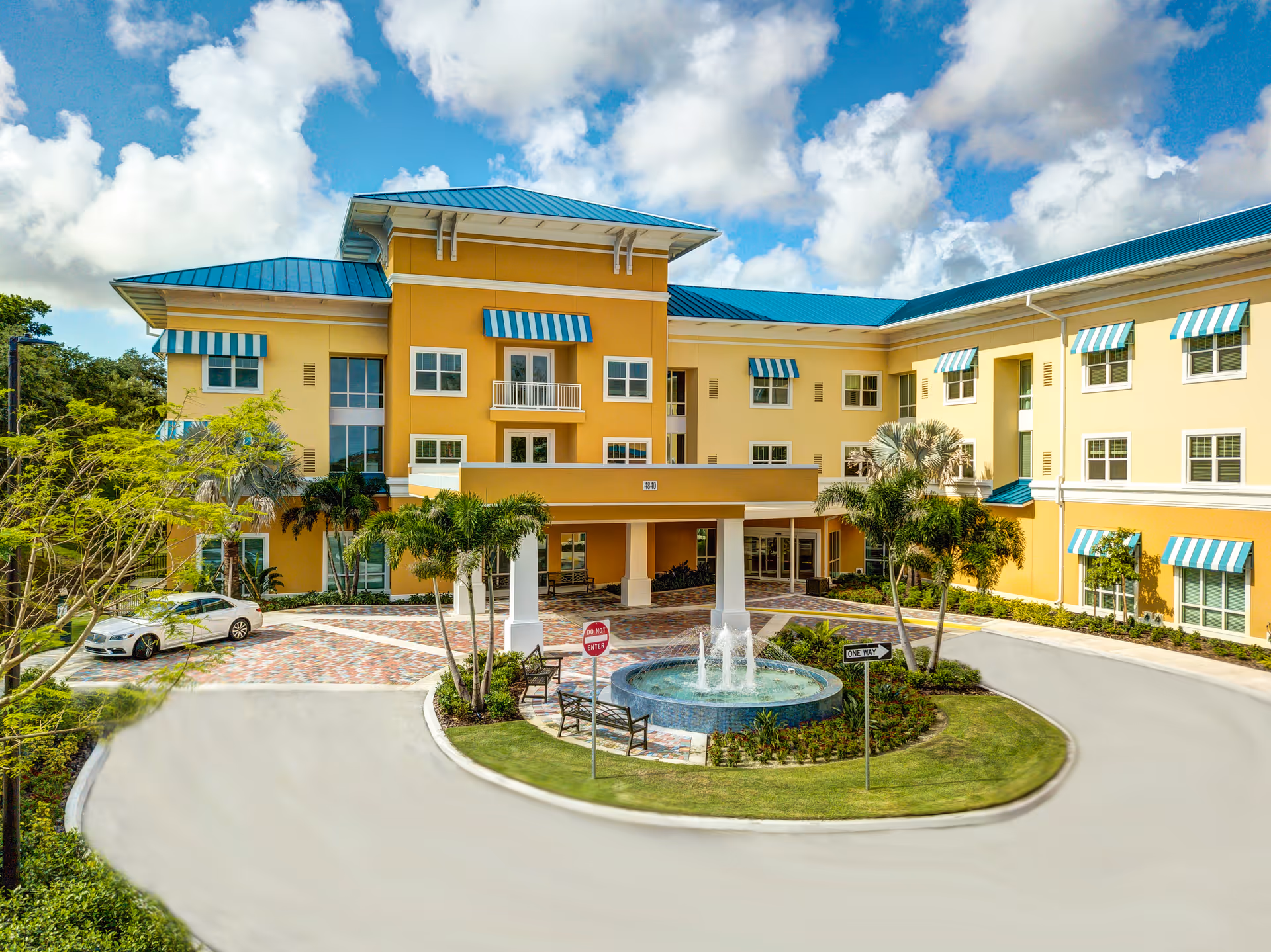 Exterior view of Symphony at Delray Beach senior living facility with a yellow building featuring blue and white striped awnings, a blue roof, a circular driveway with a fountain in the center, palm trees, and benches under a partly cloudy sky.