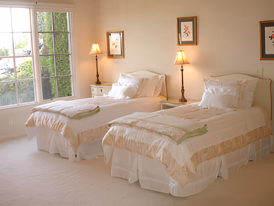 A bright bedroom with two single beds, each made with white linens and beige bed skirts. There are two bedside tables, each with a lamp, and framed artwork hanging on the wall above the beds. A large window on the left side lets in natural light and shows greenery outside.
