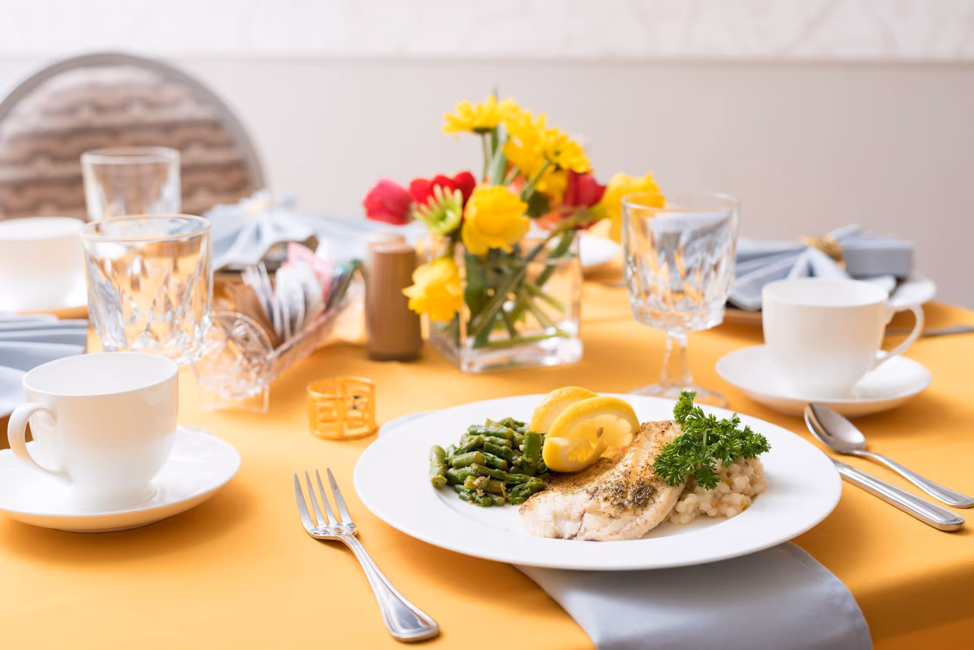 A dining table set with a plated fish entrée with asparagus and rice, coffee cups, crystal glasses, and a vase of yellow flowers on a yellow tablecloth.
