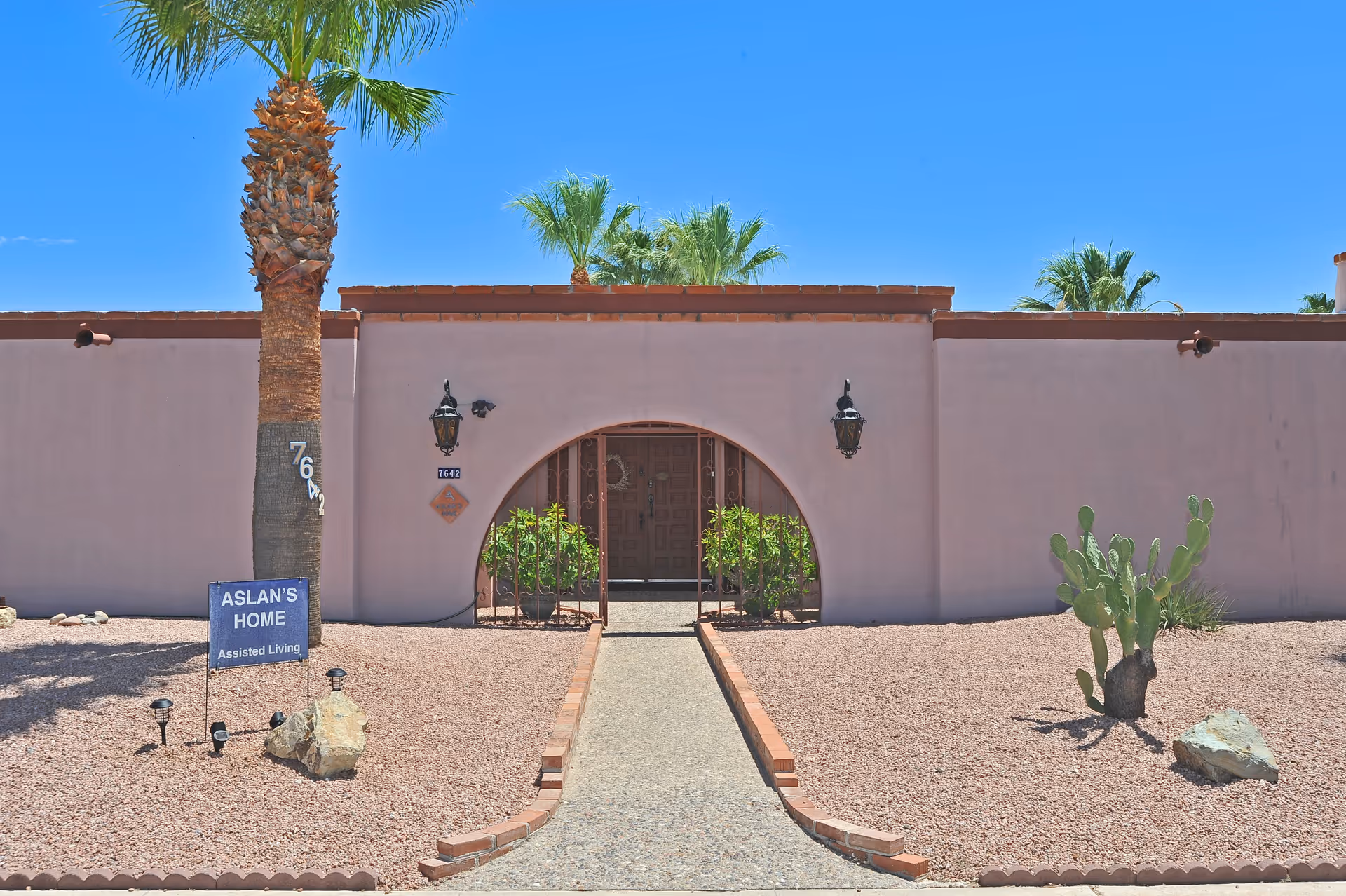 Front exterior of Aslan Assisted Living showing an arched entrance, walkway, palm trees, desert landscaping, and a sign reading "ASLAN'S HOME Assisted Living".