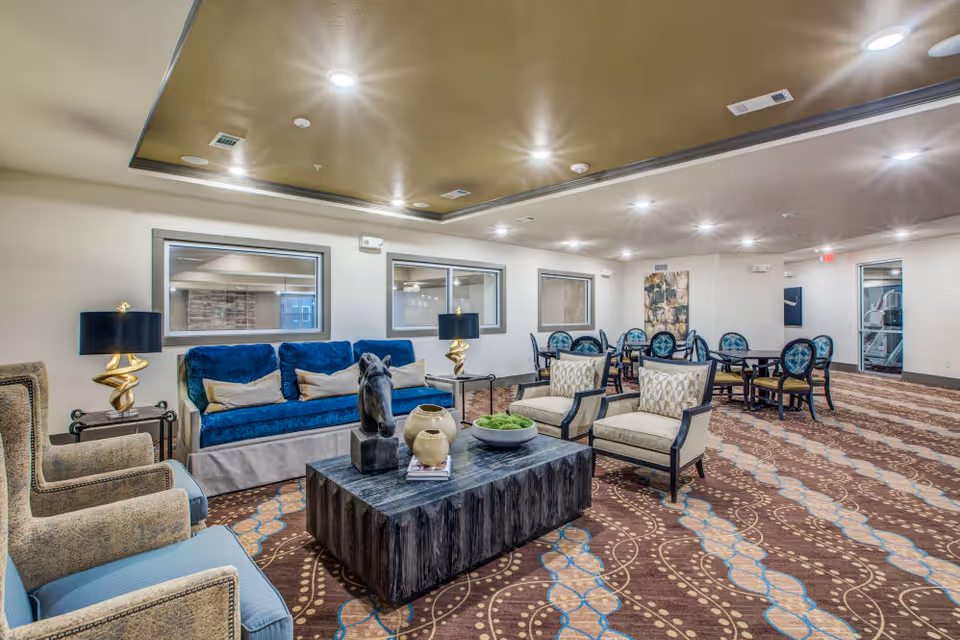 A spacious and well-lit common area in a senior living facility featuring a blue sofa with beige pillows, two beige armchairs, a dark wooden coffee table with decorative items including a horse head sculpture and vases, and several round tables with blue cushioned chairs in the background. The room has patterned carpet flooring, recessed ceiling lights, and windows on the walls.