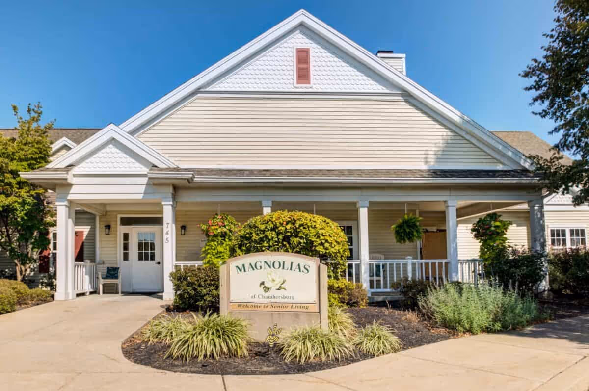 Front exterior view of a senior living facility named Magnolias of Chambersburg with a beige building, white columns, a covered porch, and a sign surrounded by greenery and plants.