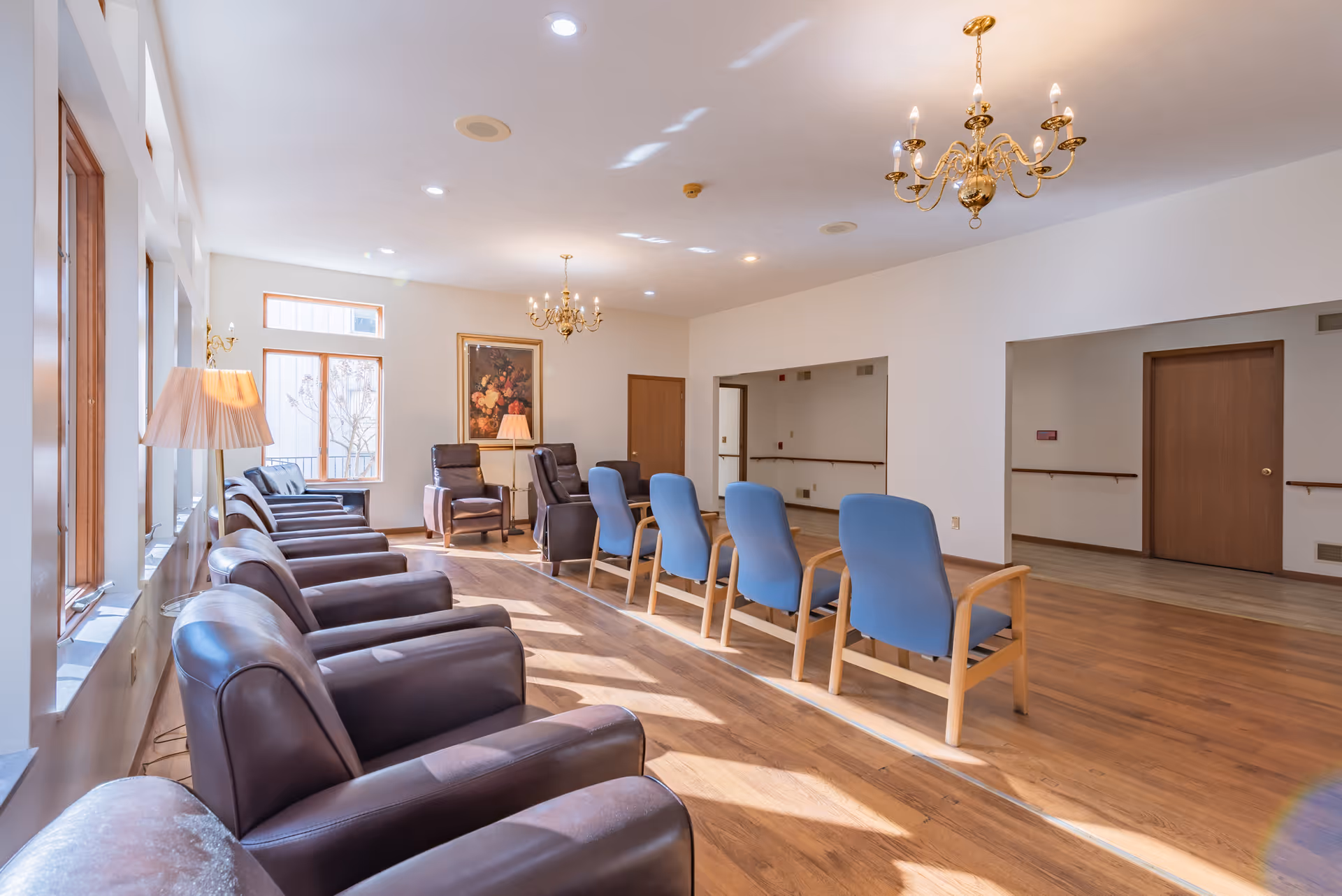 A bright and spacious common area with rows of chairs, including brown leather armchairs and blue cushioned chairs, arranged facing each other. The room features large windows letting in natural light, wooden flooring, two gold chandeliers, a floor lamp, and a framed floral painting on the wall.