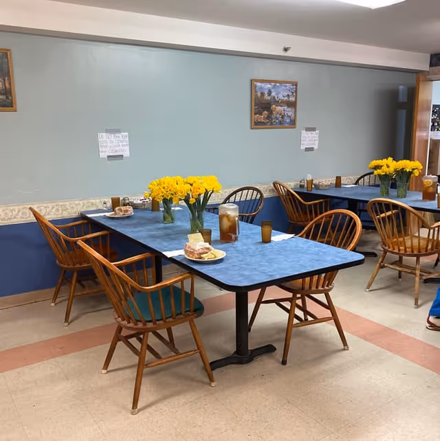 A dining area with blue tables and wooden chairs. Each table has a vase with yellow flowers and some plates with food and glasses of iced tea. The walls are painted light blue with a decorative border and have framed pictures hanging. The floor has a beige and pink tile pattern.