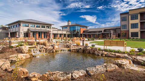 Outdoor view of Fountain View Assisted Living facility featuring a landscaped garden with a pond and small waterfalls surrounded by rocks, benches, and walking paths under a partly cloudy blue sky.