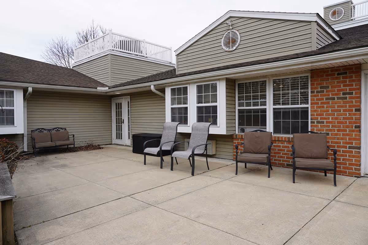 Outdoor patio area at Celebration Villa of Exeter with several cushioned chairs and a bench arranged along the building's exterior walls, which feature beige siding and red brick accents. There are multiple windows and a white door leading inside. The patio is paved with concrete and has a clean, open space.