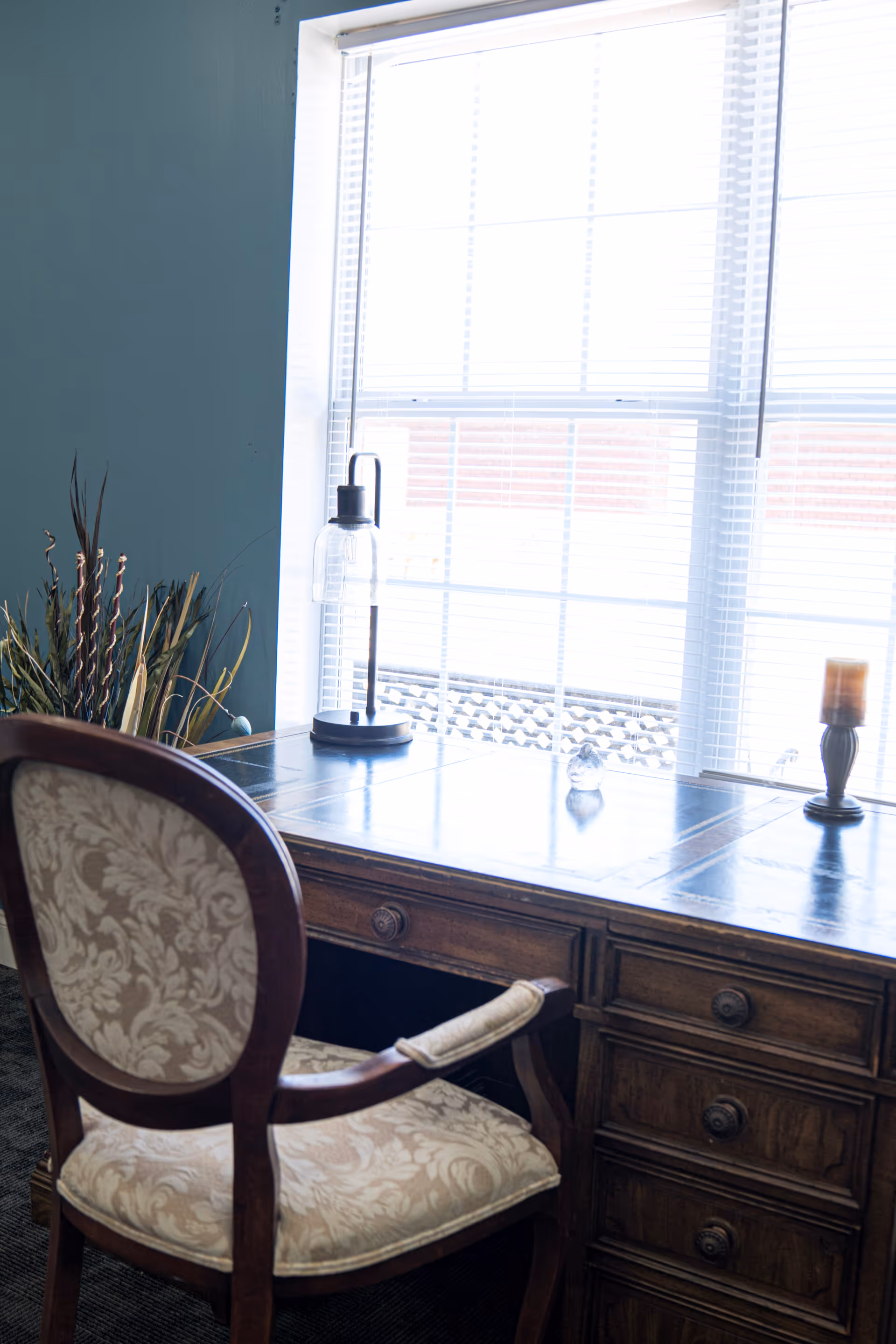 A wooden desk with multiple drawers is positioned in front of a large window with white blinds. On the desk are a modern lamp with a clear glass shade, a small glass decorative item, and a candle holder with a candle. A cushioned chair with a floral patterned fabric and wooden frame is placed in front of the desk. There is a plant with tall leaves in the corner next to the desk.
