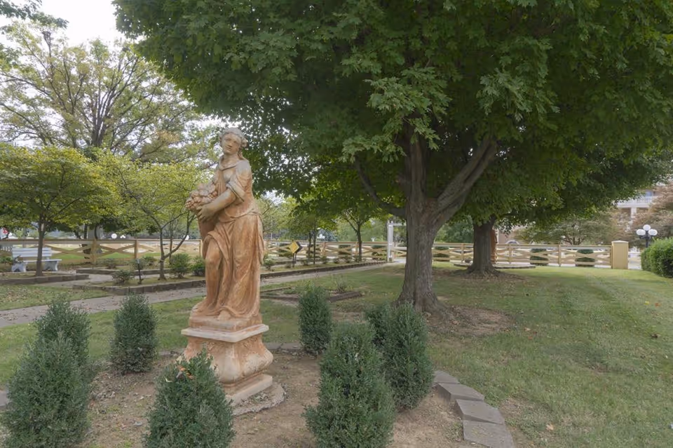 Outdoor garden area with a statue of a woman holding a basket of flowers surrounded by small bushes and large trees, with a wooden fence and benches in the background.