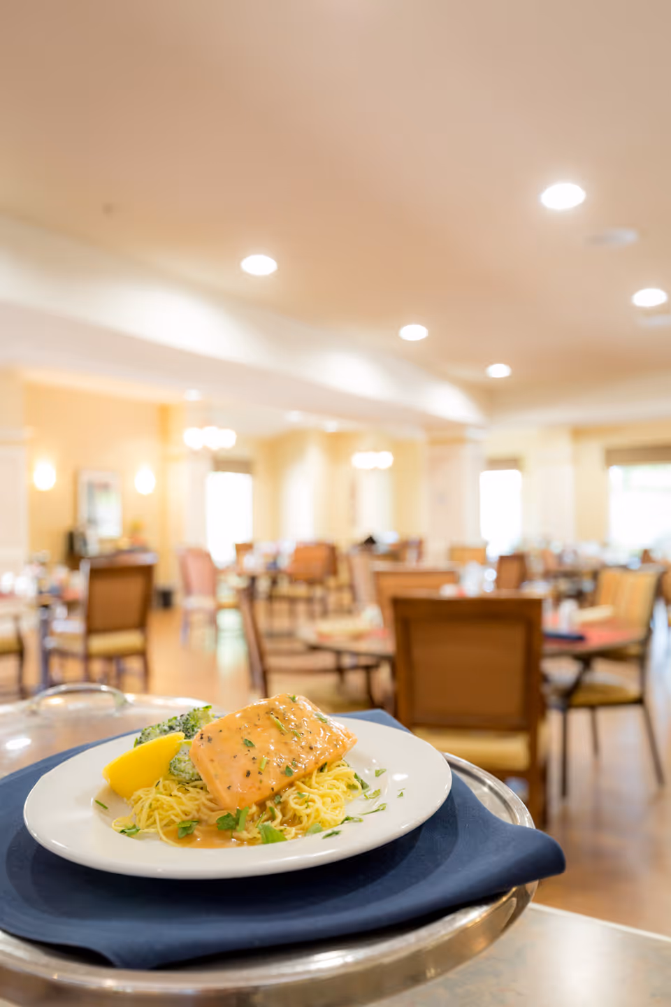 A plated meal featuring a piece of cooked salmon on a bed of noodles with a lemon wedge and broccoli, served on a tray with a navy blue napkin in a dining room with tables and chairs in the background.