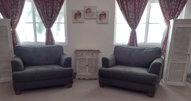 Two gray loveseats sit facing forward beneath windows with patterned red curtains and a small white cabinet between them.