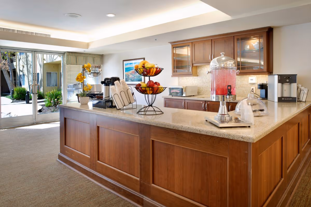 Bright communal kitchenette with a large central island holding fruit baskets, a beverage dispenser and coffee station, opening to an outdoor courtyard.