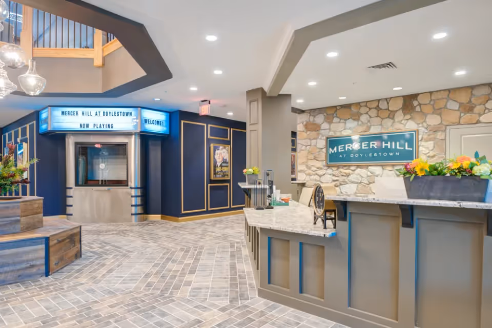 Reception area of Mercer Hill at Doylestown featuring a front desk with flowers, a stone accent wall with the facility's sign, and a hallway with a marquee sign that reads 'Mercer Hill at Doylestown Now Playing Welcome'. The space has modern lighting, tiled flooring, and decorative wall paneling.