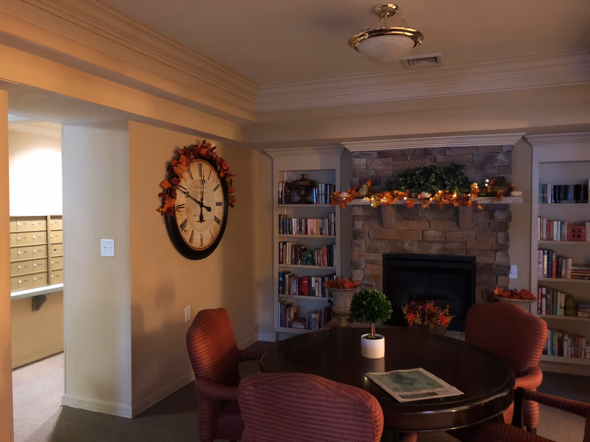 A cozy interior room with a round wooden table surrounded by four red upholstered chairs. On the table is a small potted plant and a newspaper. The room features a stone fireplace decorated with autumn-themed garlands and lights. Built-in bookshelves flank the fireplace, filled with books and decorative items. A large wall clock with Roman numerals hangs on the adjacent wall. The lighting is warm and soft, creating a welcoming atmosphere.