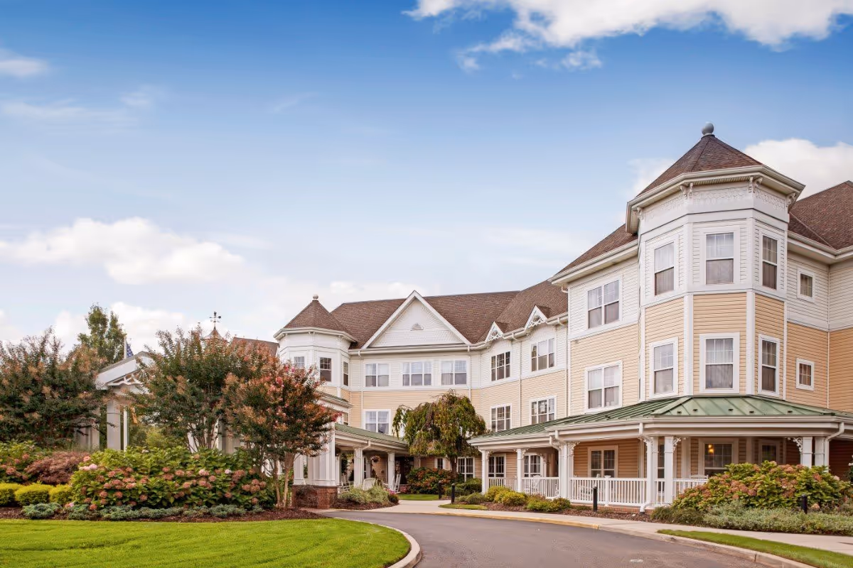 Front exterior of a large multi-story senior living building with a round turret, covered entrance, landscaped grounds and driveway under a blue sky.