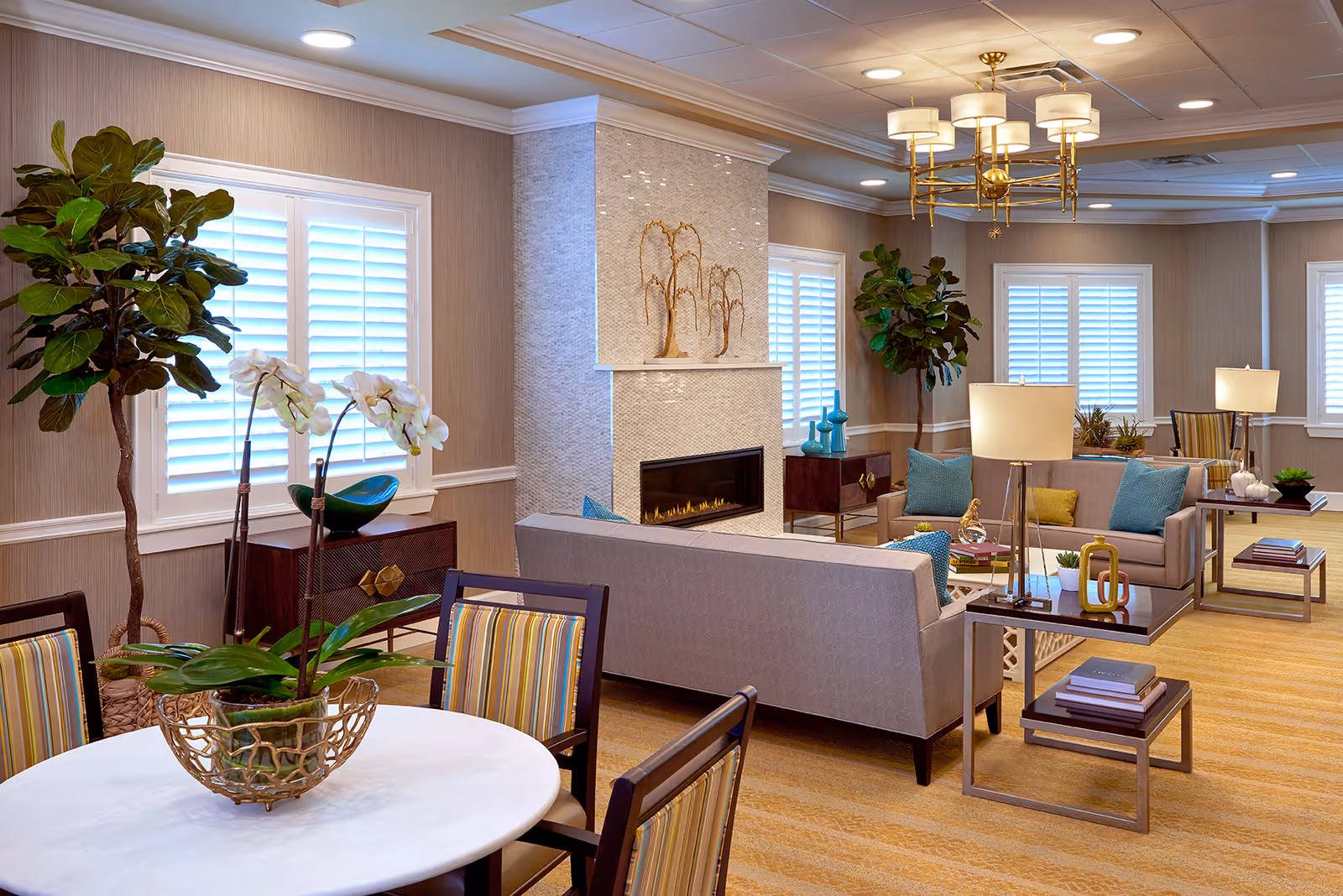 A bright and elegant living room area in an assisted living facility featuring a modern fireplace with decorative art above it, multiple seating arrangements with sofas and chairs adorned with colorful cushions, side tables with lamps and books, large windows with white shutters, indoor plants, and a round table with striped chairs and a floral centerpiece.