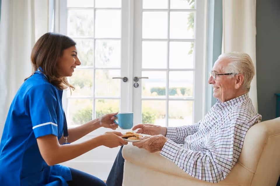 A female caregiver in a blue uniform is smiling and handing a plate of cookies and a blue mug to an elderly man with white hair and glasses who is sitting comfortably in a beige armchair near a large window with white curtains.