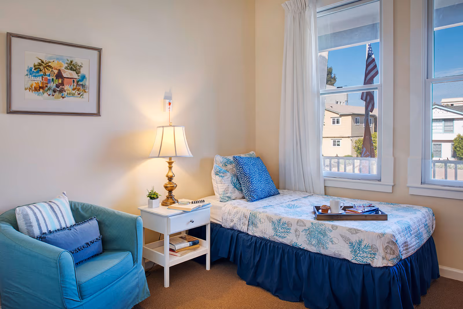 A cozy bedroom with a single bed covered in a blue and white patterned quilt and blue pillows. Next to the bed is a white nightstand with a brass lamp, a small plant, and some books. A teal armchair with two cushions is positioned to the left of the nightstand. Two windows with white curtains let in natural light and show a view of neighboring buildings and an American flag outside. A framed painting hangs on the wall above the armchair.