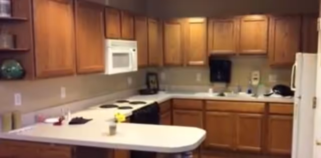 Interior view of a kitchen with wooden cabinets, a white countertop, a white microwave mounted above a stove with four burners, a refrigerator on the right, and various kitchen items on the counters.