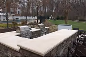 Outdoor stone bar and grilling area next to a fenced pool and lawn with trees in the background.