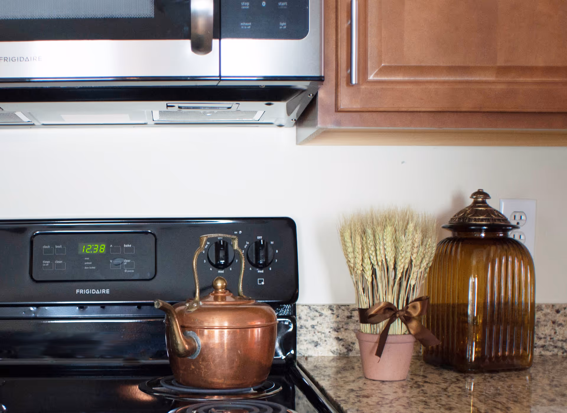 Close-up view of a kitchen stove with a copper kettle on the burner. Above the stove is a stainless steel microwave. To the right on the granite countertop, there is a small pot with wheat stalks tied with a brown ribbon and a large amber-colored glass jar with a lid. Wooden kitchen cabinets are visible above the countertop.
