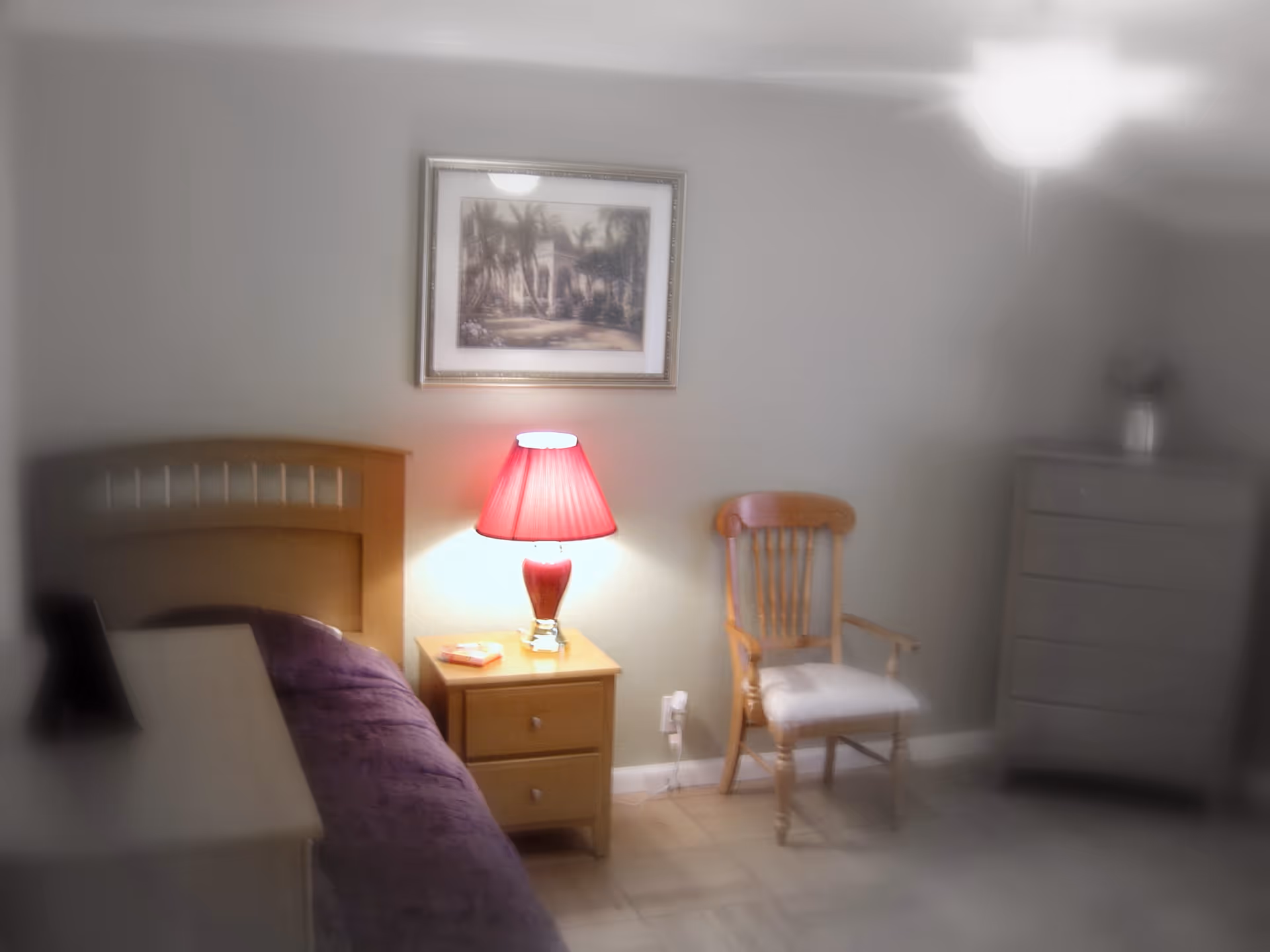 A modest bedroom with a bed, nightstand topped by a red lamp, wooden chair, framed wall art and a dresser.