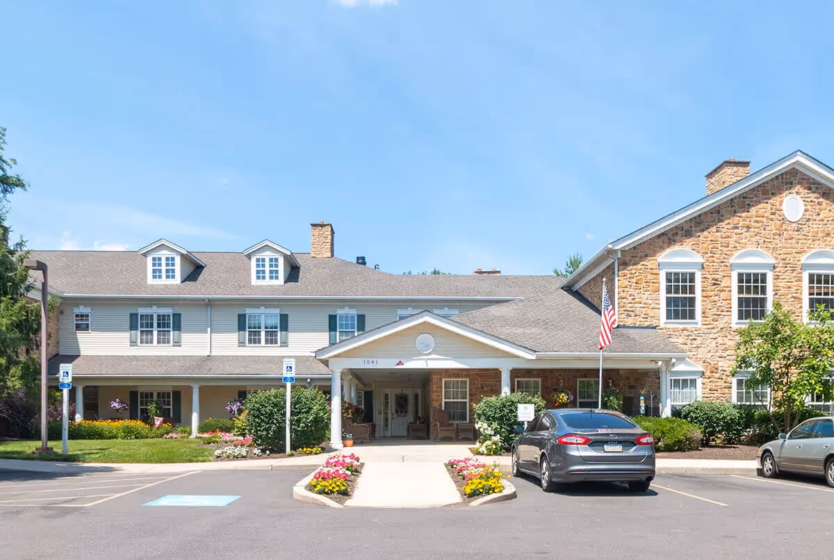 Front entrance of a two-story brick and siding senior living building with a covered porte-cochere, flowers, and parked cars.