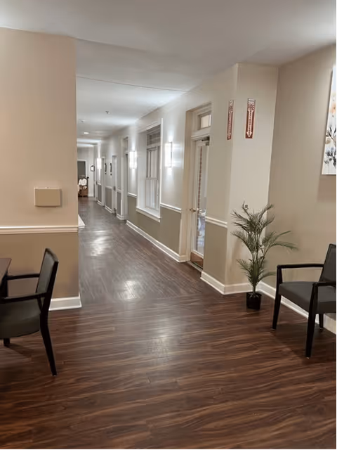 A clean, well-lit hallway in an assisted living facility with wood flooring, beige walls, and white trim. There are windows along the right side and wall-mounted lights. Two chairs and a potted plant are visible near the foreground on the right side.