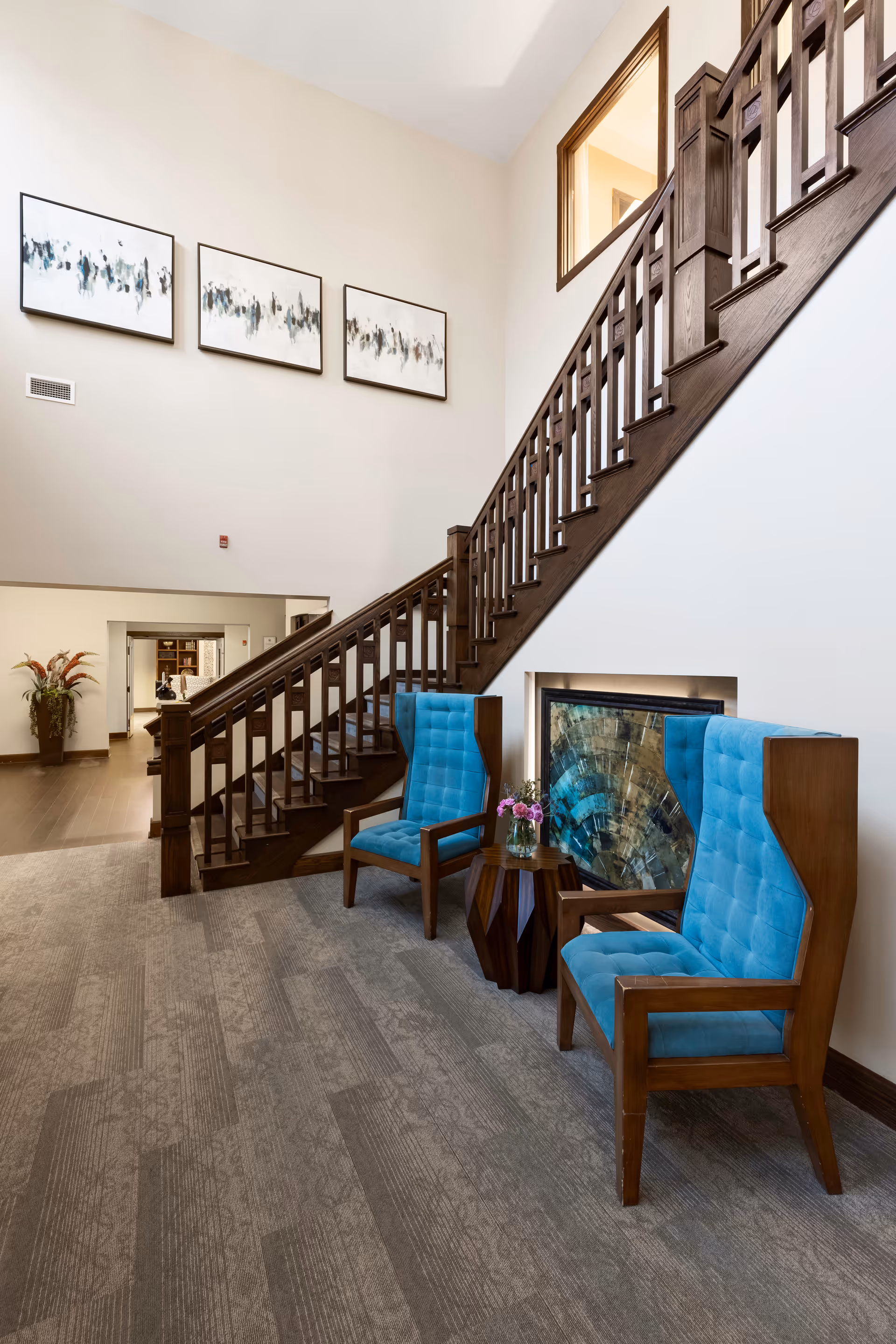 Interior view of a senior living facility featuring a wooden staircase with a dark finish. Two blue upholstered armchairs with wooden frames are placed near a small wooden side table holding a vase with flowers. There is a decorative fireplace with a stone mosaic behind the chairs. The walls are light-colored and adorned with three abstract paintings. The flooring is carpeted with a subtle pattern.