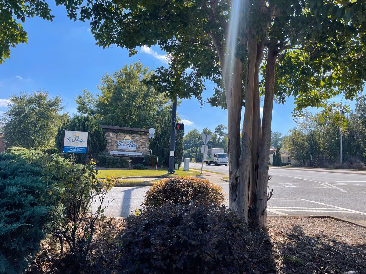 Landscaped roadside entrance with trees and a stone sign for TerraBella Sugarloaf beside a street.
