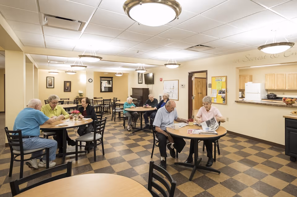Several seniors sitting at round tables in a bright communal dining/activity room with a checkered floor and a kitchen pass-through.