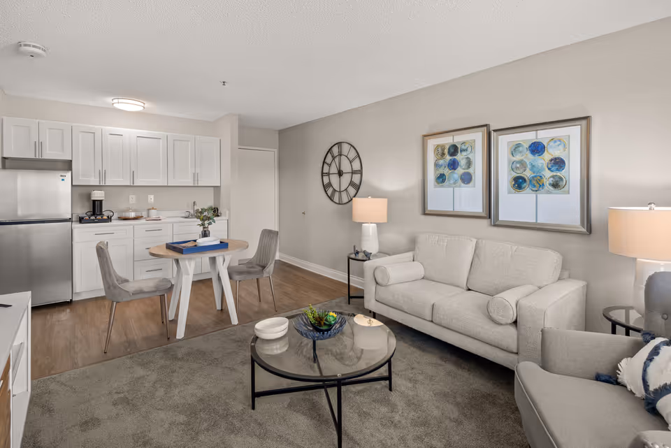 A cozy senior living community interior featuring a small kitchen area with white cabinets, a stainless steel refrigerator, and a coffee maker. Adjacent to the kitchen is a round wooden dining table with two gray upholstered chairs. The living area includes a light gray sofa, a matching armchair with a decorative pillow, a glass coffee table with decorative items, two framed abstract artworks on the wall, a large wall clock, and two table lamps providing warm lighting.
