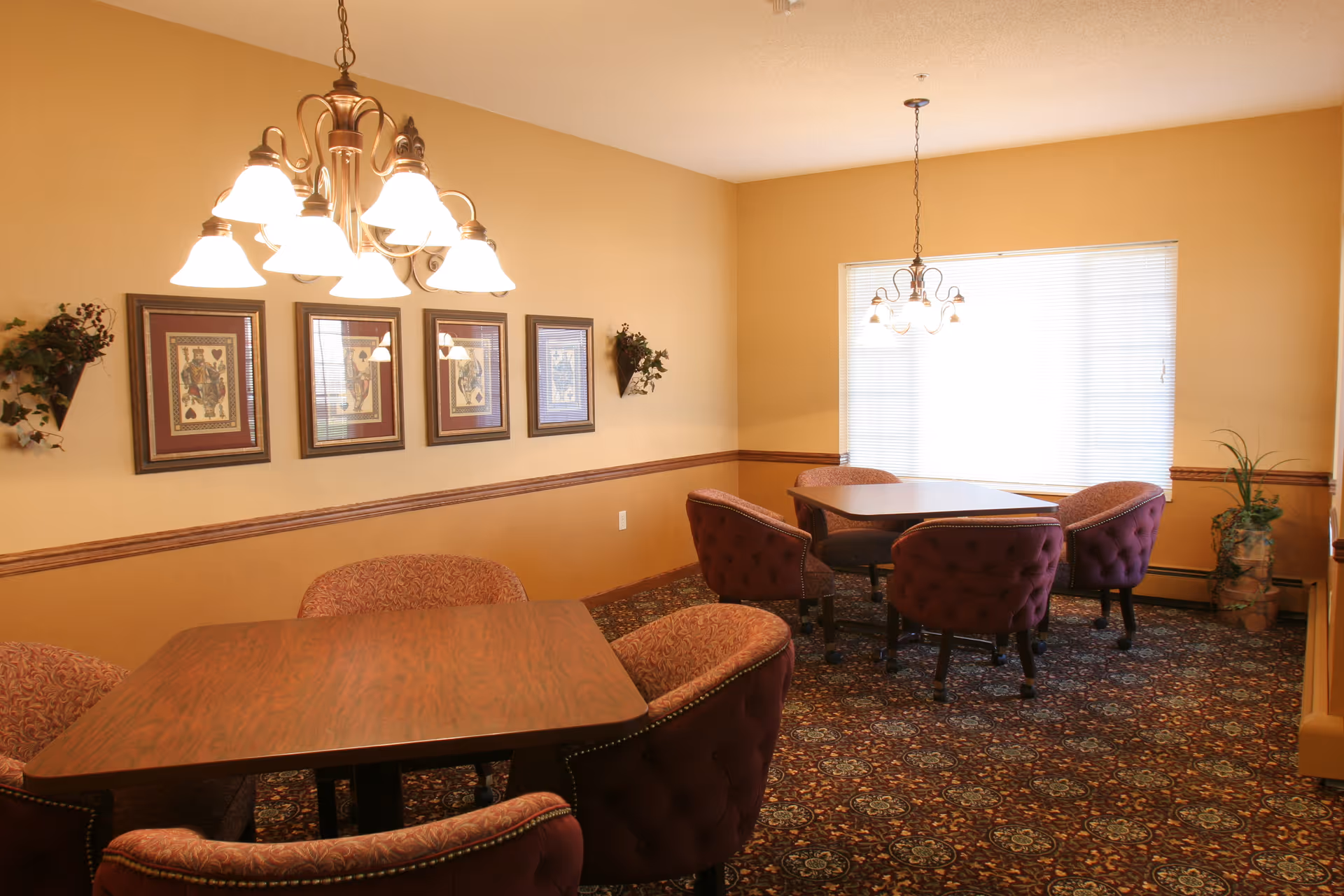Well-lit common dining room with multiple wooden tables, upholstered chairs, chandeliers, framed artwork, and patterned carpet.