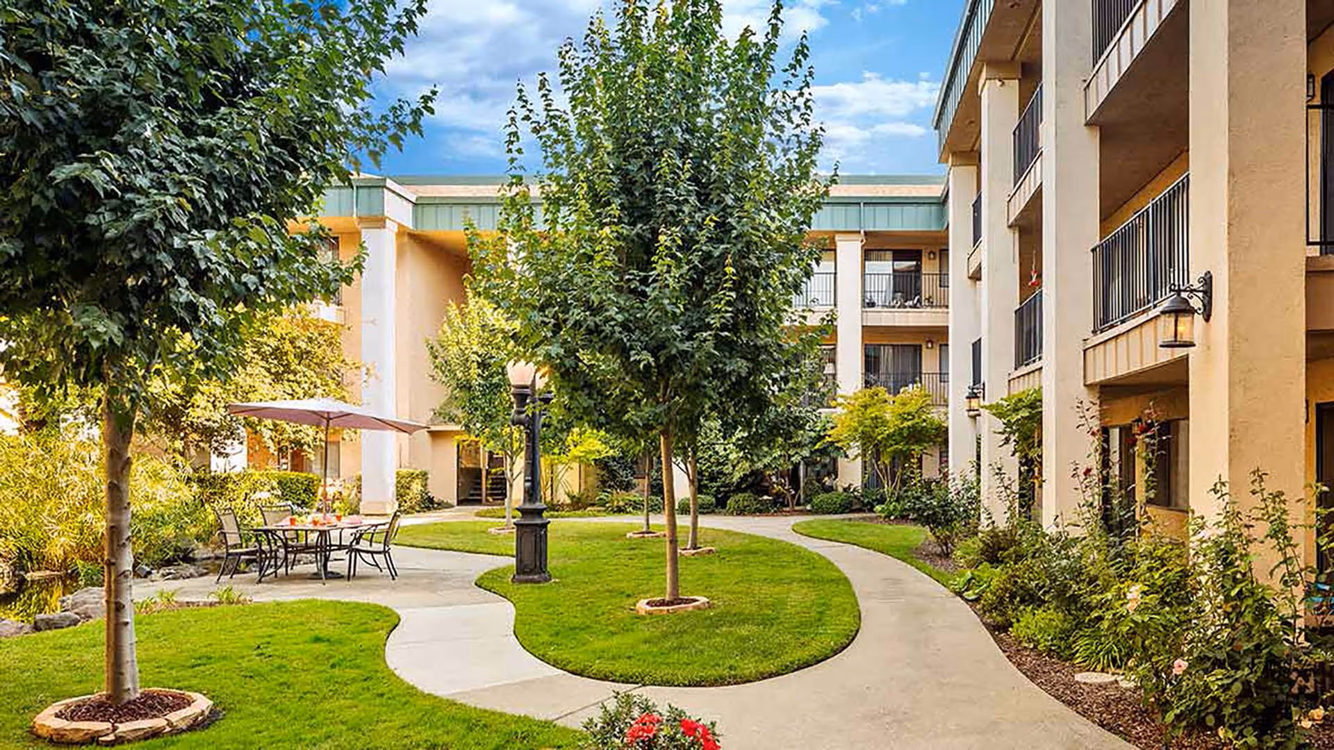 Outdoor courtyard area at a senior living facility with a curved concrete walkway, green grass, several trees, and a table with chairs under a large umbrella. The courtyard is surrounded by a multi-story beige building with balconies and windows.