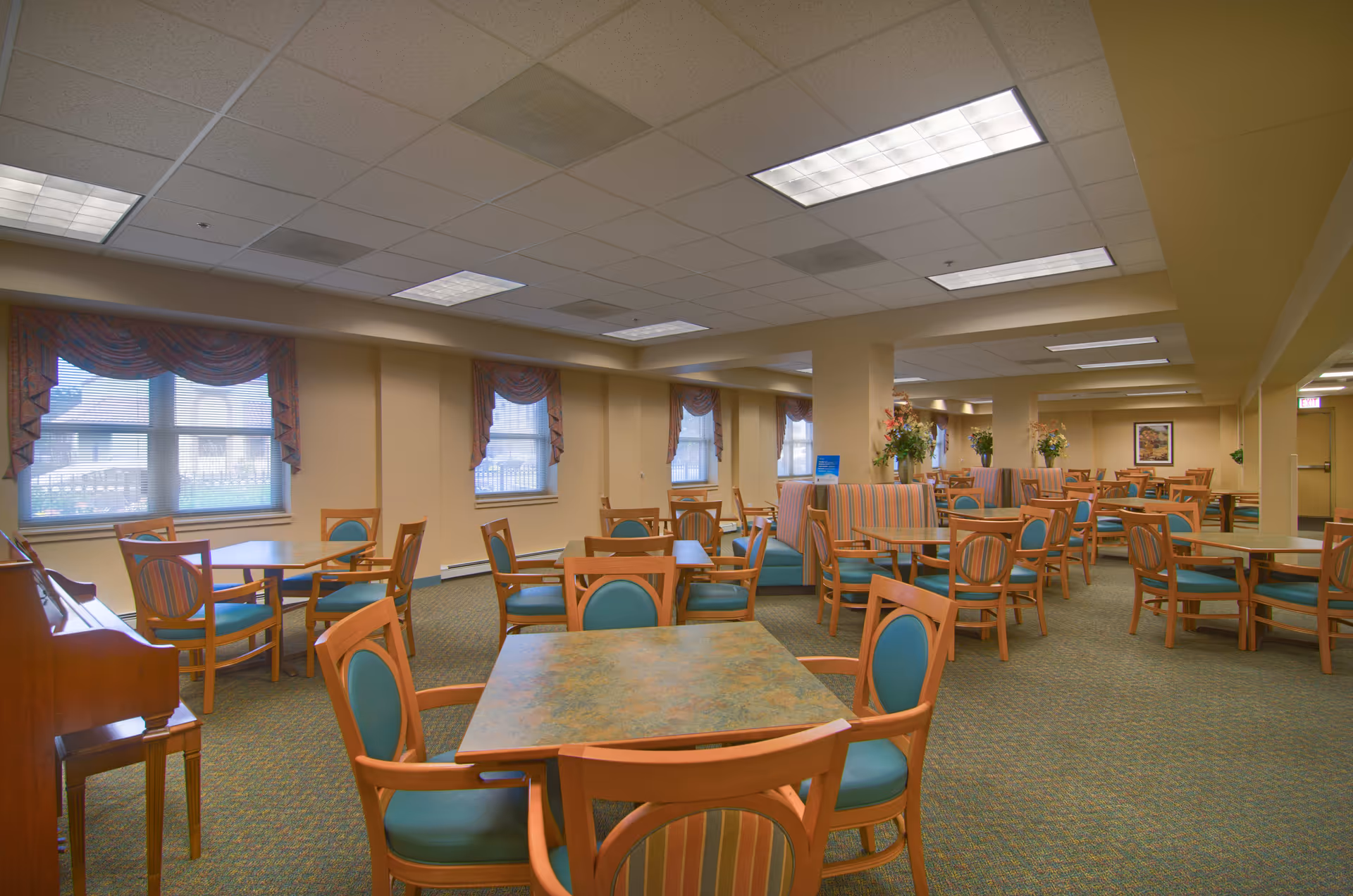 A spacious dining room in a senior living facility with multiple wooden tables and chairs featuring teal cushions. The room has beige walls, patterned carpet, and several windows with decorative valances. There are also striped upholstered booths and floral arrangements placed on some tables. The ceiling has recessed lighting panels.