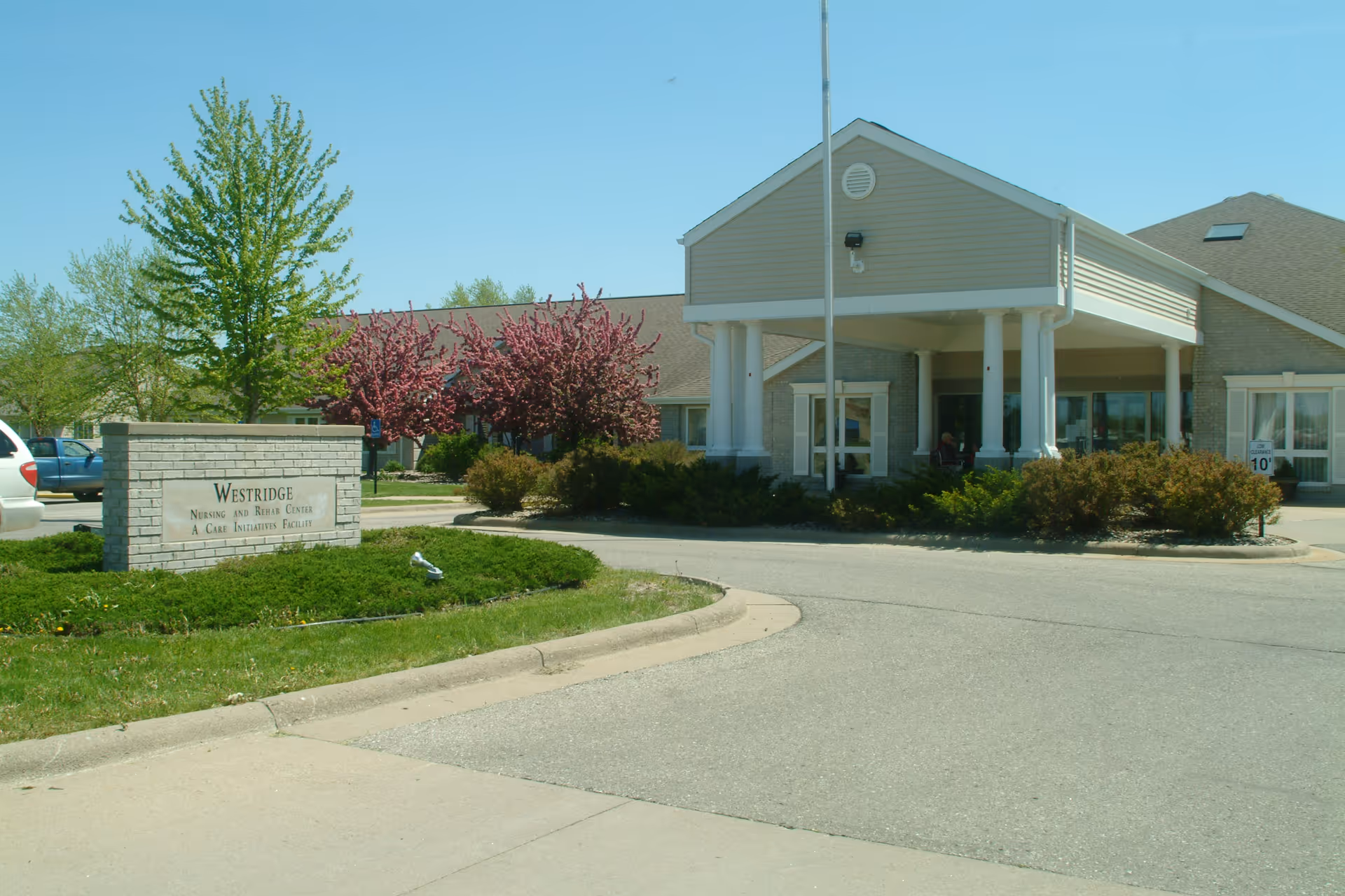 Exterior view of West Ridge Specialty Care facility showing the entrance with a covered drop-off area, surrounded by bushes and trees with a clear blue sky.