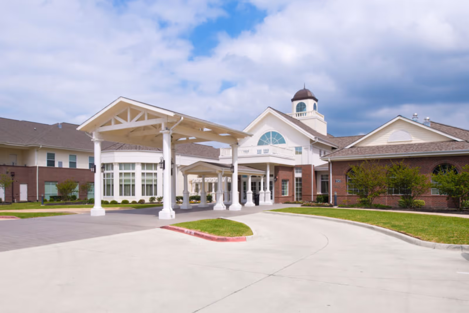 Exterior view of Crestview Retirement Community building featuring a covered entrance with white pillars, brick and white siding facade, large windows, and a cupola on the roof under a partly cloudy sky.