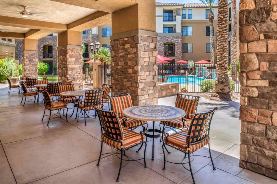 Covered outdoor patio area with round mosaic tables and striped cushioned chairs, stone pillars, and a view of a swimming pool with red umbrellas and palm trees in the background.