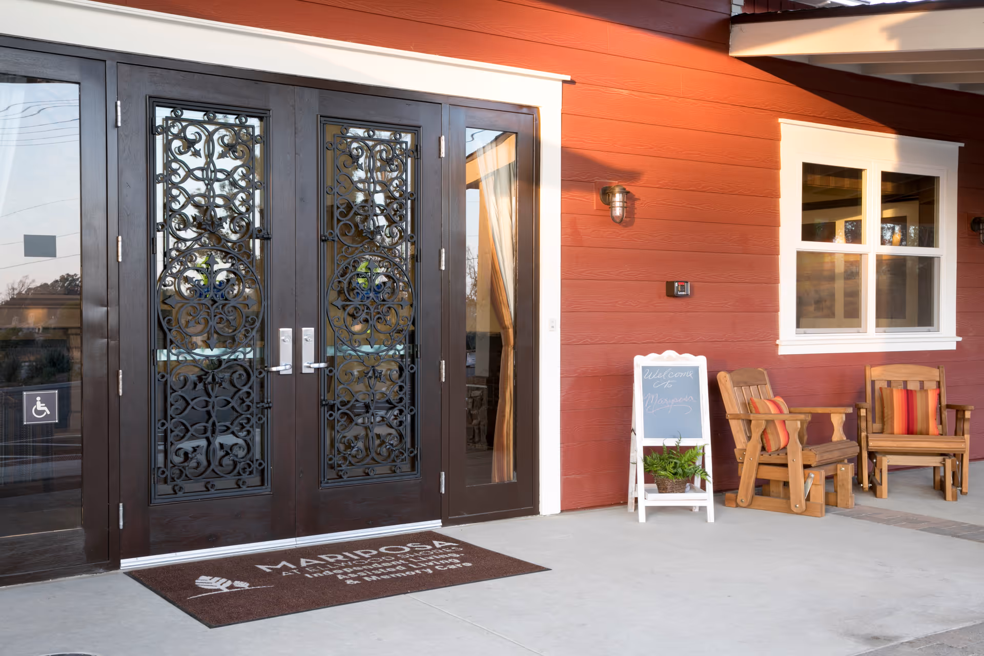 Entrance to a building with double dark wooden doors featuring intricate wrought iron designs. The exterior wall is painted red with white trim around a window and door. Two wooden chairs with red and orange striped cushions are placed on the right side next to a small white chalkboard sign with a plant underneath. A brown doormat with white text is in front of the doors.