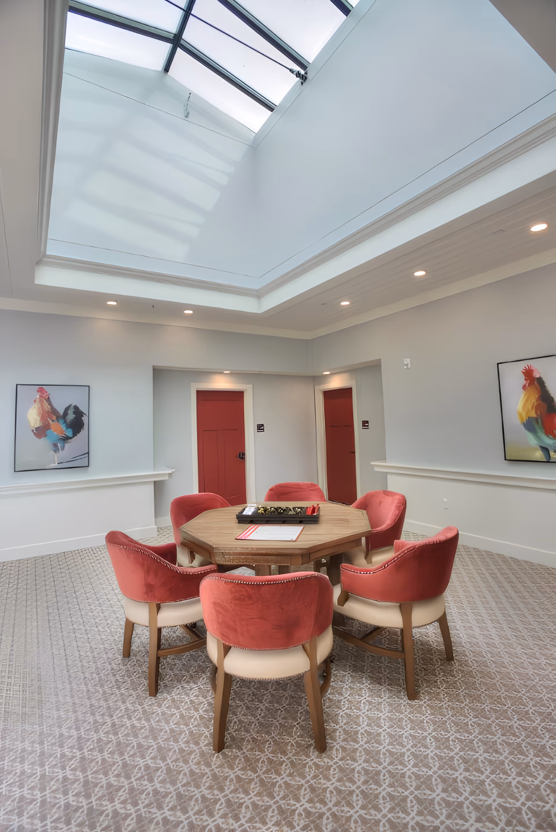 A bright room with a large skylight ceiling, featuring a round wooden table surrounded by six red and beige upholstered chairs. The walls are light gray with two red doors and two framed paintings of roosters. The floor is carpeted with a patterned design.