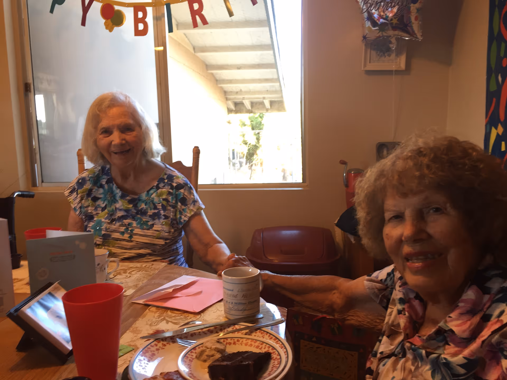 Two elderly women sitting at a dining table in a room decorated for a birthday celebration. One woman is smiling and wearing a floral top, while the other is holding her hand and also smiling. On the table are plates with cake, a red cup, a coffee mug, greeting cards, and a tablet. A 'Happy Birthday' banner is partially visible hanging near a window in the background.