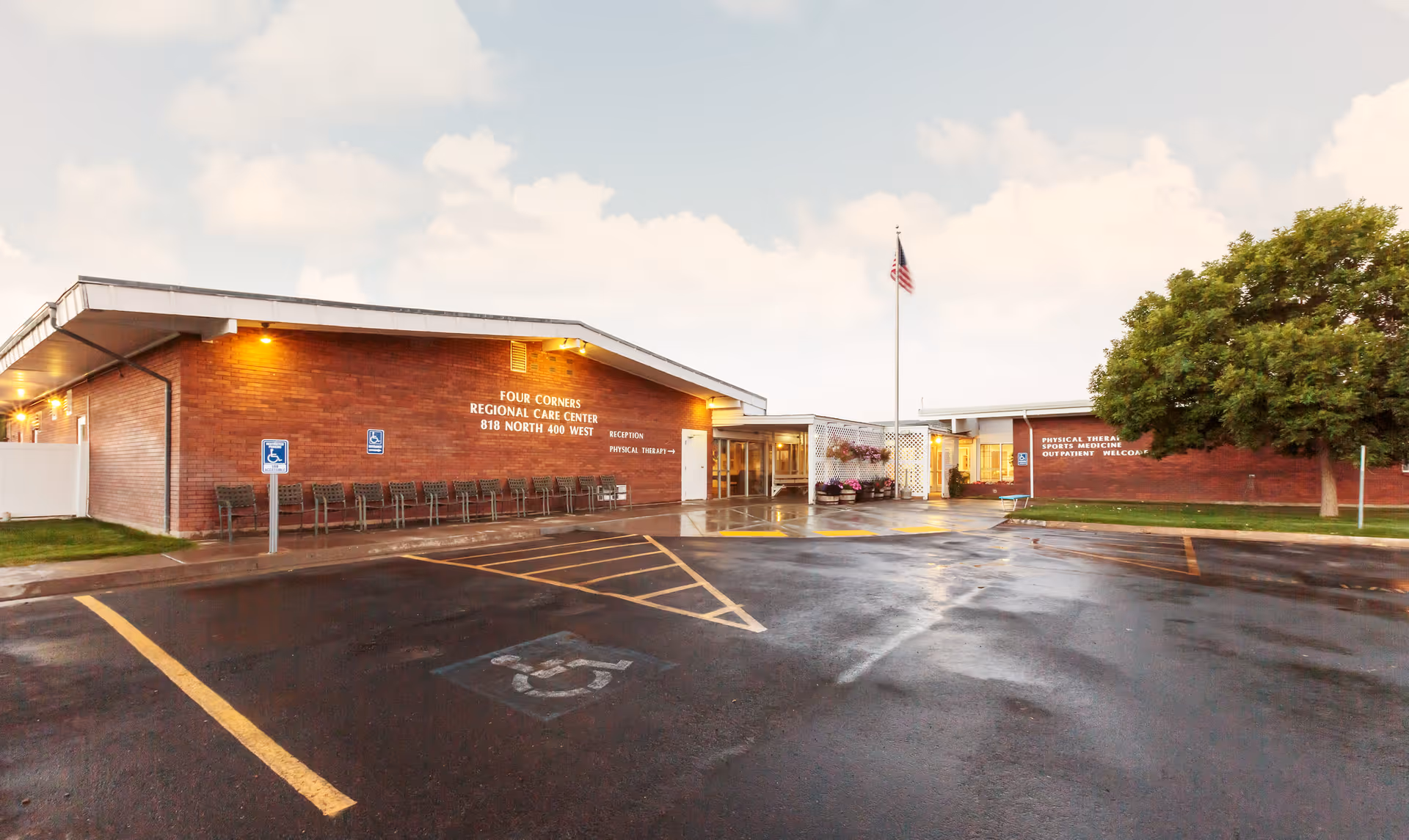 Exterior view of Four Corners Regional Care Center building with a parking lot in front, including handicap parking spaces, a flagpole with an American flag, and a large tree on the right side. The building has brick walls and illuminated signage indicating the facility name and address.