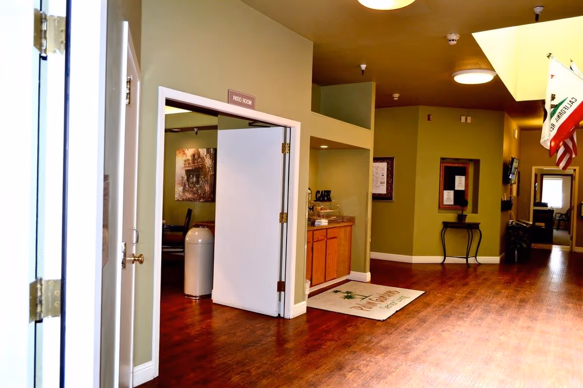 Interior hallway of Woodland Gardens Senior Living with green walls and wooden flooring. A white door labeled 'Patio Room' is open, revealing a room with chairs and a painting on the wall. There is a small counter area with a sign that says 'CAFE' and a mat on the floor with the facility's name. Flags are hanging from the ceiling on the right side of the hallway.