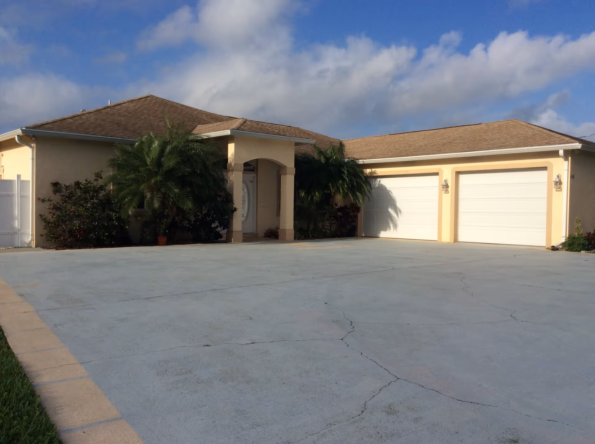 Exterior view of a single-story residential building with a beige facade, brown shingled roof, two white garage doors, and a small covered entrance. There are palm trees and shrubs near the entrance, and a large concrete driveway in front.
