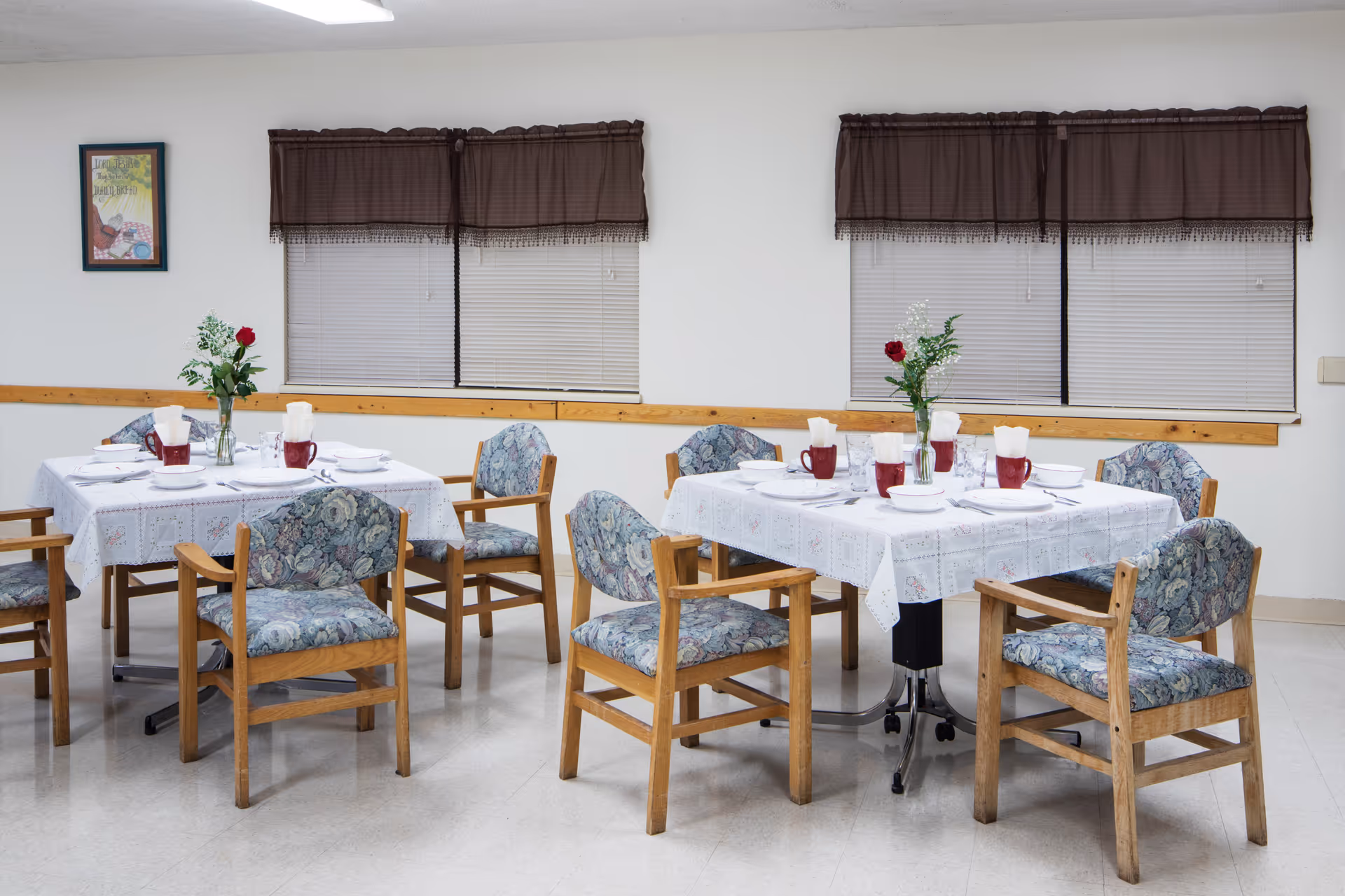A dining room with two tables covered with white tablecloths, each set with white bowls, plates, utensils, and red mugs. Each table has a vase with a single red rose and greenery. The chairs around the tables have wooden frames and floral-patterned cushions. The room has white walls, two windows with closed blinds and brown valances, and a framed picture on the wall.