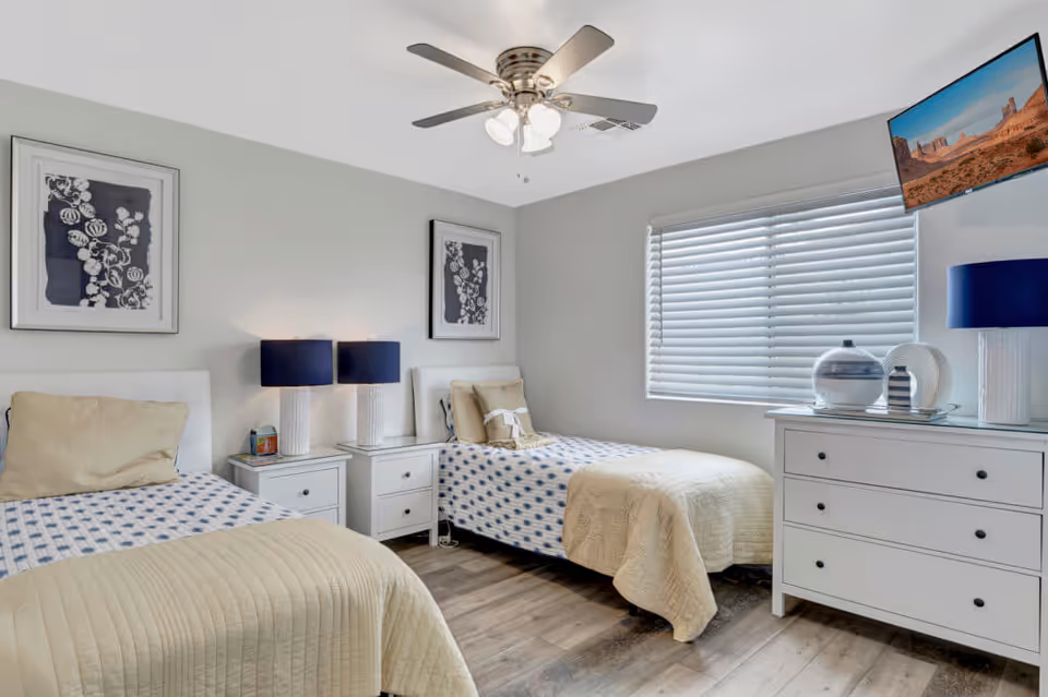 A bright bedroom with two twin beds, each with beige and blue bedding. Between the beds is a white nightstand with two matching lamps with dark blue shades. Above each bed hangs a framed black and white floral artwork. A white dresser with decorative items and a blue lamp is positioned under a window with white blinds. A ceiling fan with lights is mounted on the ceiling, and a flat-screen TV is angled in the corner near the window.