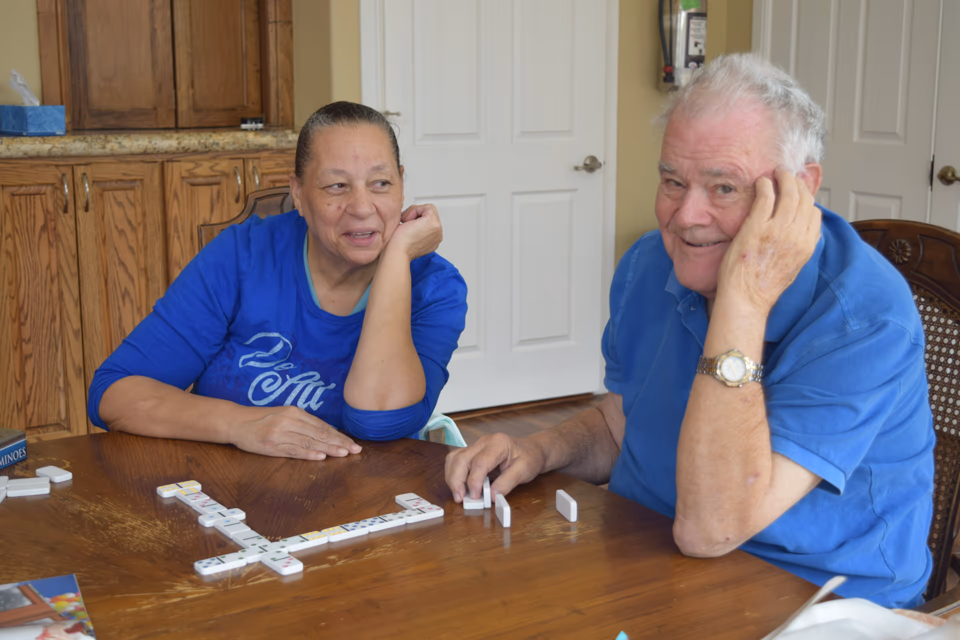 Two elderly residents seated at a wooden table indoors playing dominoes and smiling.