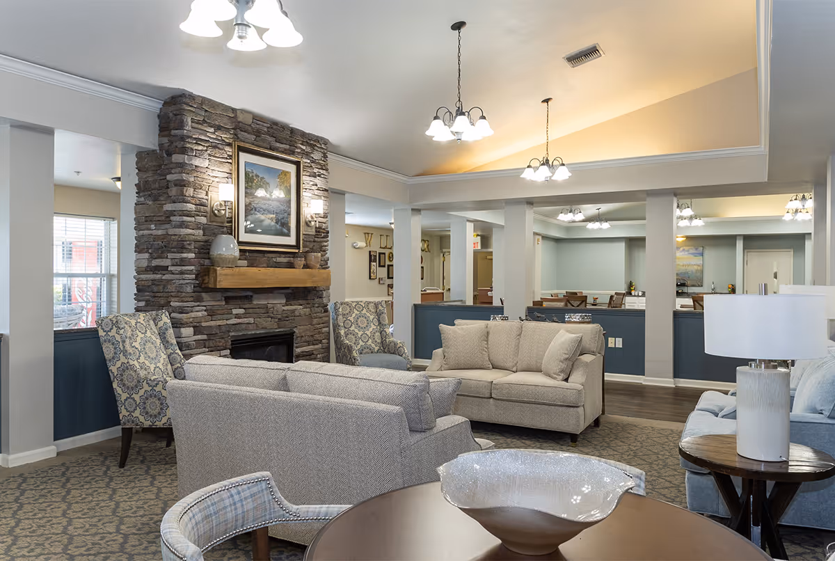 A cozy living room area in a senior living facility featuring a stone fireplace with a framed picture above it, surrounded by comfortable upholstered chairs and sofas in neutral tones. The room has soft lighting from ceiling fixtures and table lamps, with a patterned carpet and a round wooden table with a decorative bowl in the foreground.