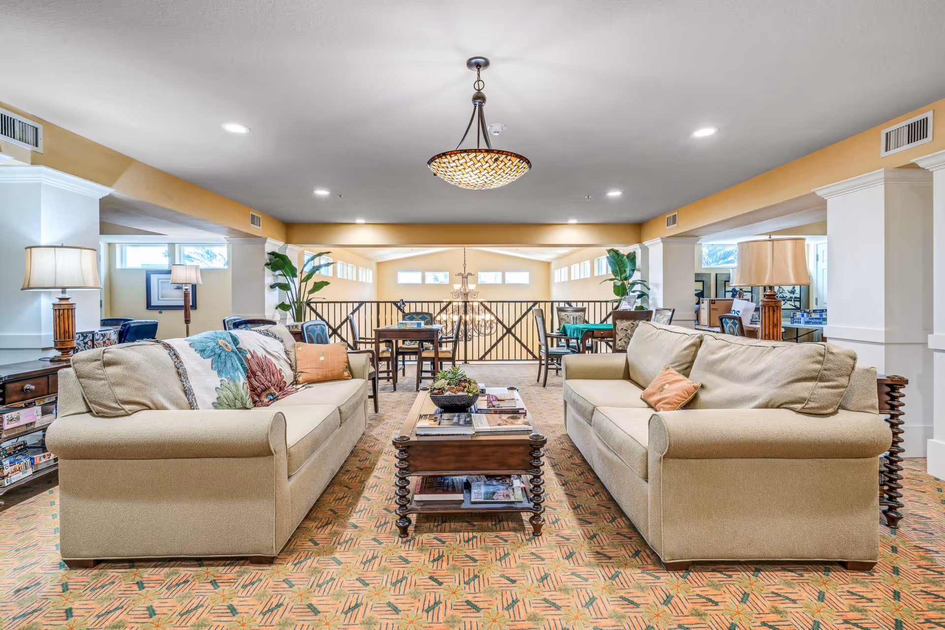 A spacious and well-lit living room area with two beige sofas facing each other, a wooden coffee table in between, and decorative pillows on the sofas. The room features a patterned carpet, table lamps on side tables, and large windows letting in natural light. In the background, there are additional seating areas and a railing overlooking a lower level with a chandelier.