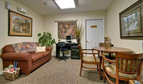 A cozy interior room with a brown couch covered by a patterned throw, a basket with knitting supplies on the floor, a small desk with a computer and office chair, a round wooden table with four chairs, framed artwork on the walls, and a potted plant near the desk.